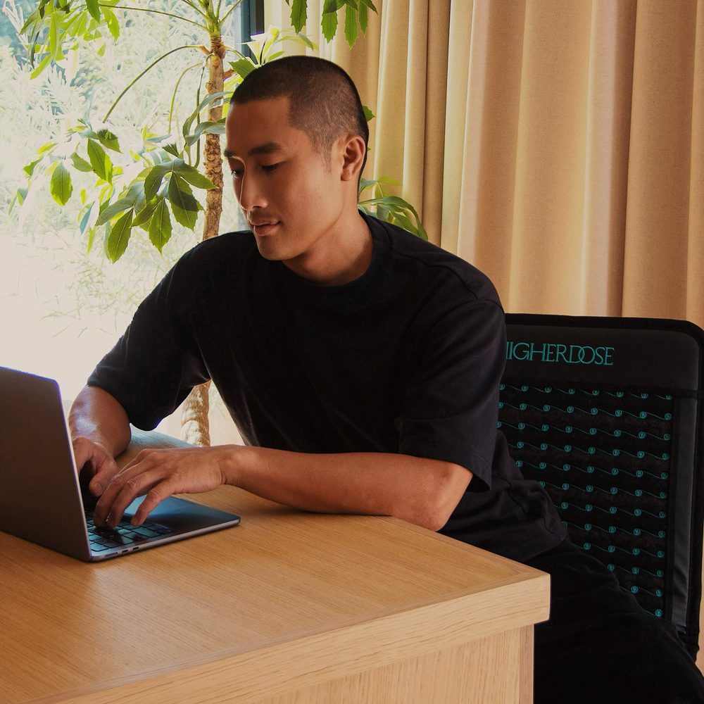 A man works at a desk while sitting on the HigherDOSE Infrared PEMF Go Mat draped over his chair.