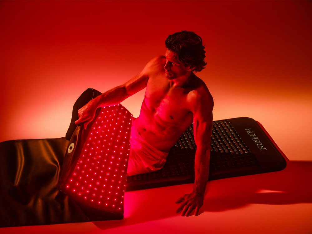 A man reclines on the HigherDOSE Infrared PEMF Pro Mat with the Full Body Red Light Mat draped over his legs.